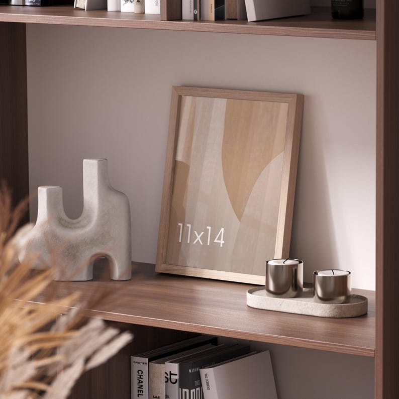 May include: A light wood picture frame with an 11x14 inch print featuring abstract beige and tan shapes. Beside it sits an irregular white ceramic sculpture and two metallic candles on an oval tray, all on a dark wood shelf.