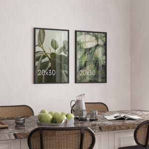 May include: Two rectangular wall frames with black borders, each displaying the text 20x30. The left frame shows green rubber plant leaves, and the right frame shows green palm fronds. The frames are mounted on a plain wall.
