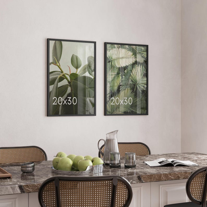 May include: Two rectangular wall frames with black borders, each displaying the text 20x30. The left frame shows green rubber plant leaves, and the right frame shows green palm fronds. The frames are mounted on a plain wall.