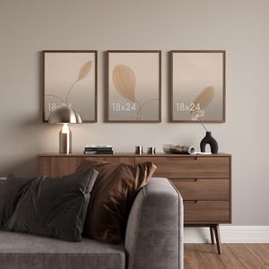 May include: Three rectangular wall frames with dark wood borders, each displaying a beige botanical print with a single leaf illustration and the white text 18x24. The frames are arranged horizontally in a row on a plain wall surface.
