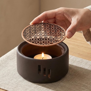 May include: A dark brown ceramic tea warmer with a decorative, openwork lid. The warmer has a cylindrical shape with vertical slots and a lit tea light inside. The lid is being lifted, revealing the tea light. The warmer sits on a neutral-coloured runner.