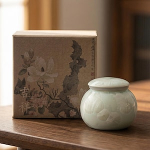 May include: A small, round, pale green ceramic jar with a lid, featuring a floral design. Beside it is a cardboard box with a floral illustration in shades of brown and white. The jar and box are on a wooden surface.