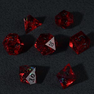 Op de afbeelding: Set van zeven rode polyhedrale dobbelstenen voor tabletopspellen. Elke dobbelsteen heeft een andere vorm, met witte cijfers en een glinsterend, doorschijnend uiterlijk. De dobbelstenen liggen op een donkergrijze ondergrond.