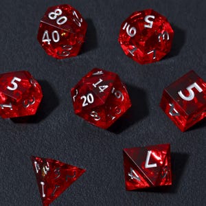 Pode incluir: Conjunto de sete dados poliédricos vermelhos com numeração branca. Os dados têm várias formas, incluindo um d4, d6, d8, d10, d12, d20 e um dado percentual. Os dados são translúcidos e têm uma aparência marmorizada.