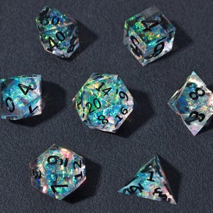 May include: Set of seven iridescent polyhedral dice with black numbering. The dice are clear with a shimmering, colorful interior, reflecting blues, greens, and purples. The dice are arranged on a dark gray surface.