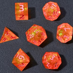 May include: Set of seven orange and yellow polyhedral dice with pink numbering. The dice are transparent and have a glittery interior. Includes a d4, d6, d8, d10, d12, d20, and a cube.