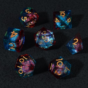Puede incluir: Un conjunto de siete dados poliédricos de varias formas, incluyendo un d4, d6, d8, d10, d12, d20 y un dado de porcentaje. Los dados son transparentes con un esquema de color azul y rojo, y numeración dorada.