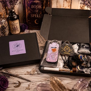 Pode incluir: Uma caixa de presente de papelão preta contendo uma coleção de itens, incluindo um chaveiro rosa e roxo com tema celestial, uma planchette preta em forma de coração com texto dourado, um pequeno frasco de vidro e vários cristais.