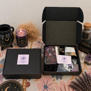 Op de afbeelding: Twee zwarte dozen, waarvan één open, gevuld met diverse items, waaronder tarotkaarten, stickers en copalhars. Een brandende paarse kaars en een decoratieve mok zijn zichtbaar. De dozen hebben de tekst "Witchy Wonders Box" erop.