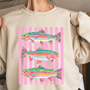 May include: A beige crewneck sweatshirt featuring a graphic print of three colorful rainbow trout fish arranged horizontally. The fish are layered over a background of vertical light pink stripes, showcasing vibrant hues of blue, green, and pink.