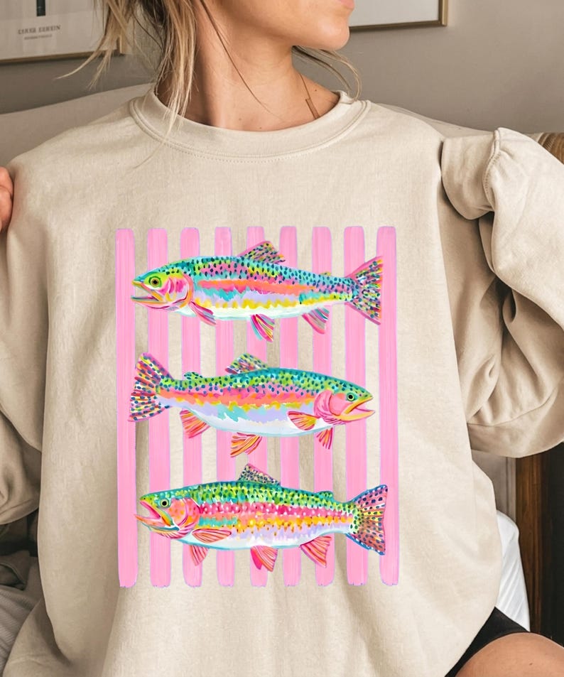 May include: A beige crewneck sweatshirt featuring a graphic print of three colorful rainbow trout fish arranged horizontally. The fish are layered over a background of vertical light pink stripes, showcasing vibrant hues of blue, green, and pink.
