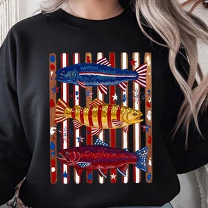 May include: A black crewneck sweatshirt featuring a graphic print of three fish layered over vertical red, white, and blue stripes. The top fish is blue, the middle is yellow, and the bottom is red, all accented with star and firework motifs.