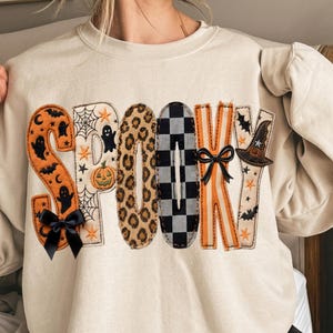 Op de afbeelding: Een beige sweater met ronde hals en het woord SPOOKY in grote, decoratieve letters. Elke letter heeft een uniek patroon, waaronder oranje met spoken, wit met spinnenwebben en pompoenen, luipaardprint, zwart-wit geblokt en oranje strepen.