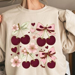 May include: A beige crewneck sweatshirt featuring a graphic print of dark red cherries, light pink cherry blossoms, and pink bows with gold outlines. The design is scattered with small dark red and light pink heart shapes across the front.
