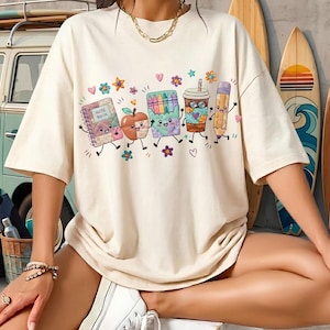 May include: A cream-colored t-shirt featuring a graphic print of a notebook, an apple, a box of crayons, a coffee cup, and a pencil, all with smiling faces and limbs, surrounded by small colorful flowers and hearts on the front chest.
