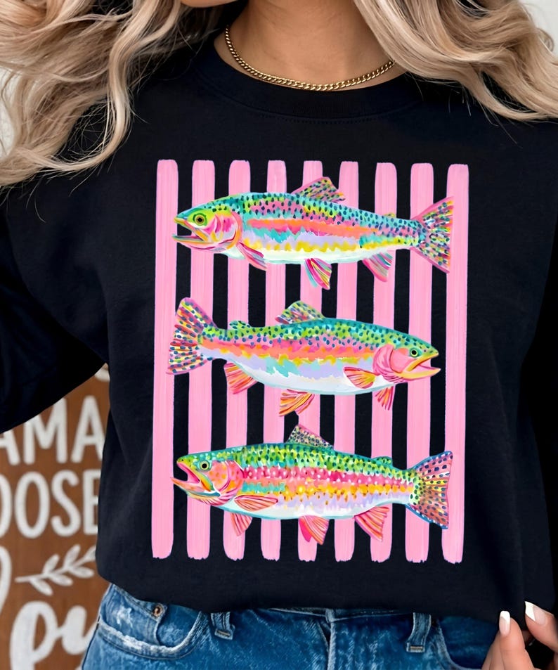 May include: A black long-sleeve sweatshirt featuring a graphic print of three colorful rainbow trout fish layered over a background of vertical pink and black stripes. The fish are depicted in vibrant shades of green, pink, yellow, and blue.