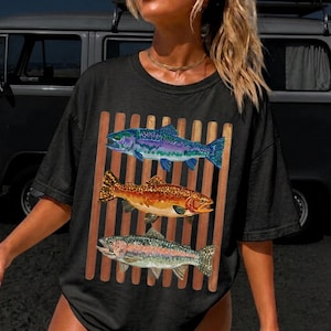 May include: A black short-sleeve t-shirt featuring a graphic print of three fish stacked vertically. The top fish is blue and purple, the middle fish is orange and brown with spots, and the bottom fish is silver and pink. The fish are set against brown stripes.