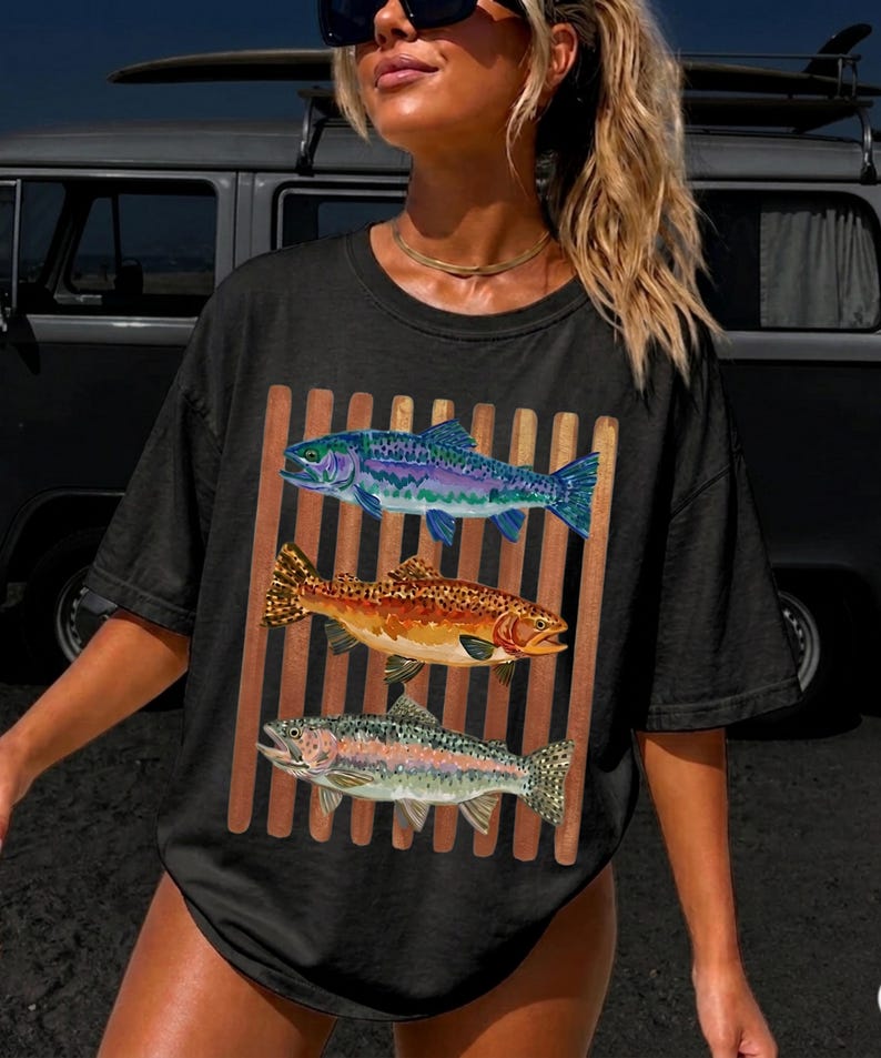 May include: A black short-sleeve t-shirt featuring a graphic print of three fish stacked vertically. The top fish is blue and purple, the middle fish is orange and brown with spots, and the bottom fish is silver and pink. The fish are set against brown stripes.