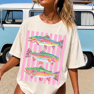 May include: An off-white short-sleeve t-shirt featuring a graphic print of three colorful rainbow trout fish arranged vertically. The fish are layered over a background of vertical light pink stripes, creating a vibrant and distinct visual contrast.