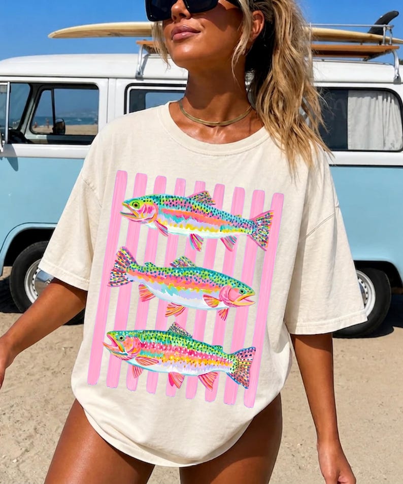 May include: An off-white short-sleeve t-shirt featuring a graphic print of three colorful rainbow trout fish arranged vertically. The fish are layered over a background of vertical light pink stripes, creating a vibrant and distinct visual contrast.