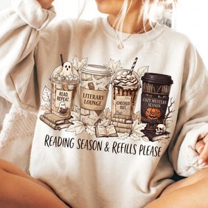 Puede incluir: Una sudadera beige de cuello redondo con un estampado gráfico de cuatro tazas de café rodeadas de hojas de otoño y libros. Las tazas tienen las etiquetas Read. Repeat., Literary Lounge, Checked Out y Cozy Mystery Season. El texto Reading Season & Refills Please aparece impreso debajo del gráfico.