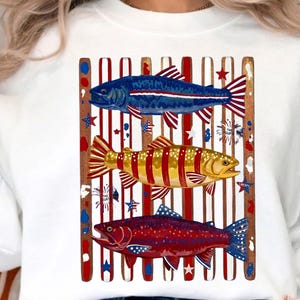 May include: A white crewneck sweatshirt featuring a graphic of three fish layered over vertical red and white stripes. The top fish is blue, the middle is yellow, and the bottom is red, all decorated with stars and stripes patterns.