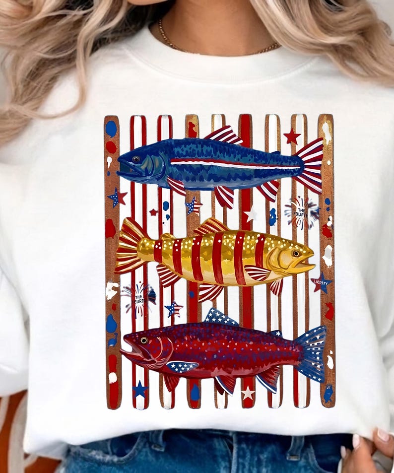 May include: A white crewneck sweatshirt featuring a graphic of three fish layered over vertical red and white stripes. The top fish is blue, the middle is yellow, and the bottom is red, all decorated with stars and stripes patterns.