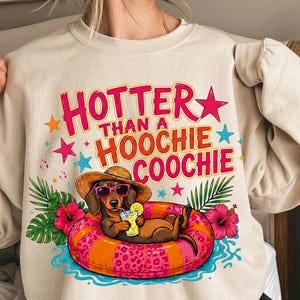 May include: Beige sweatshirt with the text "Hotter Than a Hoochie Coochie". The graphic features a dachshund wearing sunglasses and a straw hat, relaxing in a pink and orange inner tube, holding a drink. Tropical flowers and leaves surround the dog.