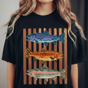 May include: A black t-shirt featuring a graphic print of three fish stacked vertically over a background of brown vertical bars. The top fish is blue and purple, the middle fish is orange and brown, and the bottom fish is silver and green.