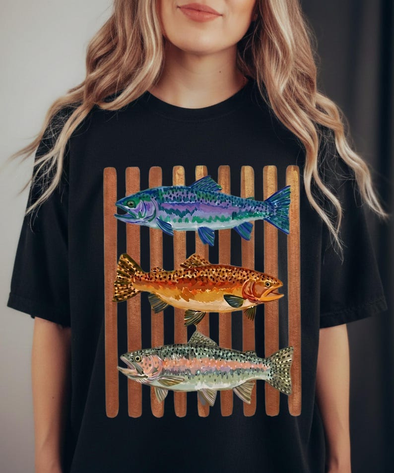 May include: A black t-shirt featuring a graphic print of three fish stacked vertically over a background of brown vertical bars. The top fish is blue and purple, the middle fish is orange and brown, and the bottom fish is silver and green.