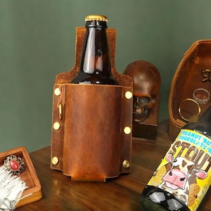 Op de afbeelding: Een bruine leren bierfleshouder met messing klinknagels en een goudkleurige sluiting. Een donkere glazen fles zit in de houder. De houder staat op een houten oppervlak.