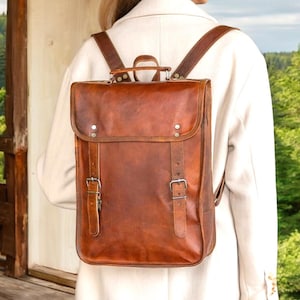 May include: A brown leather backpack with adjustable shoulder straps and a top handle. The bag has a rectangular shape with a flap closure secured by two buckles. The leather has a distressed look. The backpack is being worn.