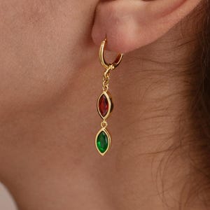 Peut inclure: Boucles d'oreilles créoles dorées avec un design pendant. Chaque boucle d'oreille présente une pierre précieuse rouge et verte de forme ovale. Les boucles d'oreilles sont portées à l'oreille d'une personne.