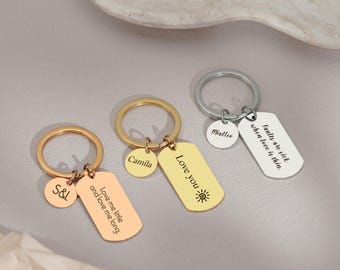 Op de afbeelding: Drie gepersonaliseerde sleutelhangers met gegraveerde labels. Eén gouden sleutelhanger met "Love you" en een zon. Een andere gouden sleutelhanger met "S&L" en "Love me little and love me long". De derde is zilverkleurig met "Miallee" en "faults are sick when love is thin."