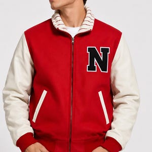 May include: A red varsity jacket with white sleeves and a ribbed collar featuring thin red stripes. The jacket has a front zipper closure, two white-trimmed pockets, and a large black letter N with a white border on the left chest.