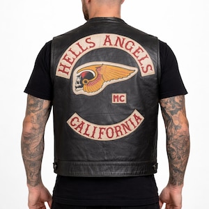 Puede incluir: Chaleco de cuero negro con el parche del logo de Hells Angels MC en la espalda. El parche presenta una calavera alada y las palabras "Hells Angels" y "California". El chaleco se lleva sobre una camiseta negra.