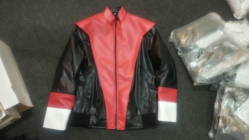 May include: A black jacket featuring a red V-shaped panel on the front, a high collar, and a full-length front zipper. The sleeves have red and white bands at the cuffs, and the bottom hem is finished with a black ribbed waistband.