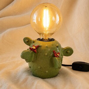 May include: A green ceramic cactus-shaped table lamp with a glossy finish, featuring small raised dots and two red flower accents. The lamp is topped with a large, round, warm-glowing Edison-style light bulb and includes a black power cord with a switch.