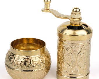 Pode incluir: Um moedor de especiarias e uma pequena tigela a condizer, dourados. O moedor tem uma manivela e um corpo cilíndrico com um padrão floral em relevo. A tigela é redonda com um design em relevo semelhante. Ambos os itens são de metal.