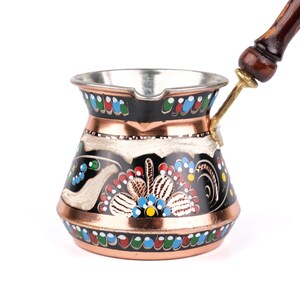 Könnte beinhalten: Eine türkische Kaffeekanne aus Kupfer mit einem dekorativen Blumendesign in Rot, Blau, Gelb und Grün auf schwarzem Hintergrund. Die Kanne hat einen Holzgriff und einen silberfarbenen Innenraum.