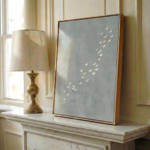 May include: A framed artwork featuring a flock of white birds in flight against a muted blue-gray background. The artwork has a gold-colored frame and is leaning against a wall. A gold-colored lamp with a beige lampshade is in the background.