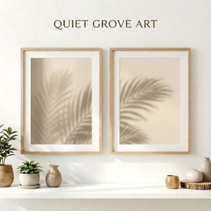May include: Two framed prints with a neutral colour palette. Each print features a beige background with the shadow of palm leaves. The frames are light wood. The text "QUIET GROVE ART" is above the frames. The prints are displayed on a white shelf.
