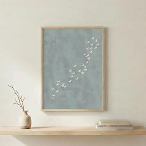 May include: A framed art print featuring a flock of white birds in flight against a muted blue-grey background. The artwork is displayed in a light wood frame. A small vase with a branch and books are on a wooden shelf below.