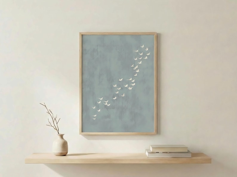 May include: A framed art print featuring a flock of white birds in flight against a muted blue-grey background. The artwork is displayed in a light wood frame. A small vase with a branch and books are on a wooden shelf below.