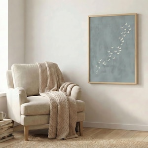 May include: A light beige armchair with a matching knitted throw. A framed artwork with a light blue background and white bird silhouettes hangs on the wall. The room has a neutral color palette with a wooden floor and a small side table.