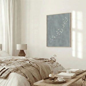 May include: A bedroom scene with a neutral color palette. A bed with a beige quilt and pillows is in the foreground. A framed artwork with a blue background and white shapes hangs on the wall. A wooden table holds books and a coffee cup.