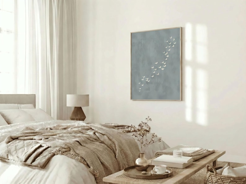 May include: A bedroom scene with a neutral color palette. A bed with a beige quilt and pillows is in the foreground. A framed artwork with a blue background and white shapes hangs on the wall. A wooden table holds books and a coffee cup.