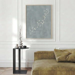 May include: A framed artwork features a muted blue background with a flock of white birds in flight. The artwork hangs above a modern black side table with a decorative vase and a sofa with olive green cushions.
