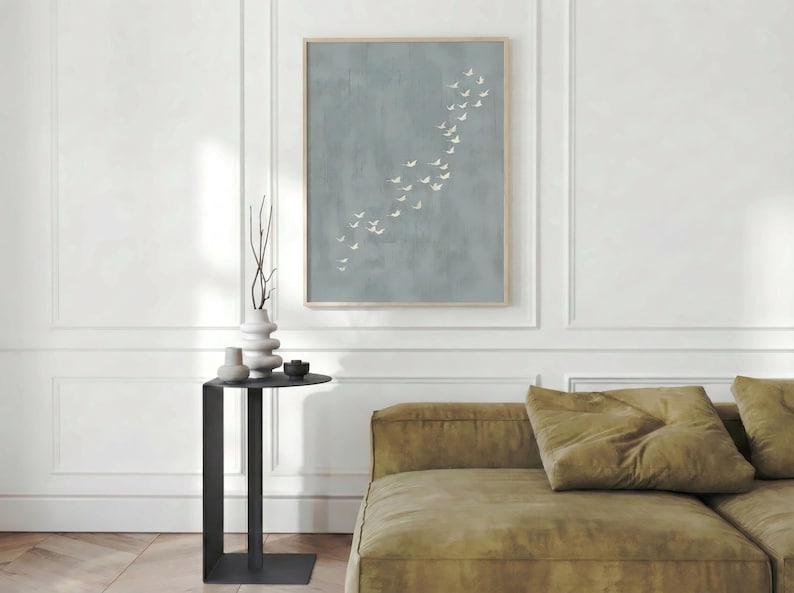 May include: A framed artwork features a muted blue background with a flock of white birds in flight. The artwork hangs above a modern black side table with a decorative vase and a sofa with olive green cushions.