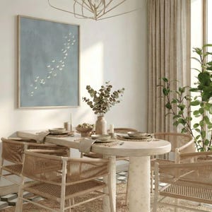 May include: A dining room scene featuring a round table with woven chairs. A framed artwork with a blue background and white bird silhouettes hangs on the wall. A vase of dried flowers and place settings adorn the table. Natural light streams in from a large window.
